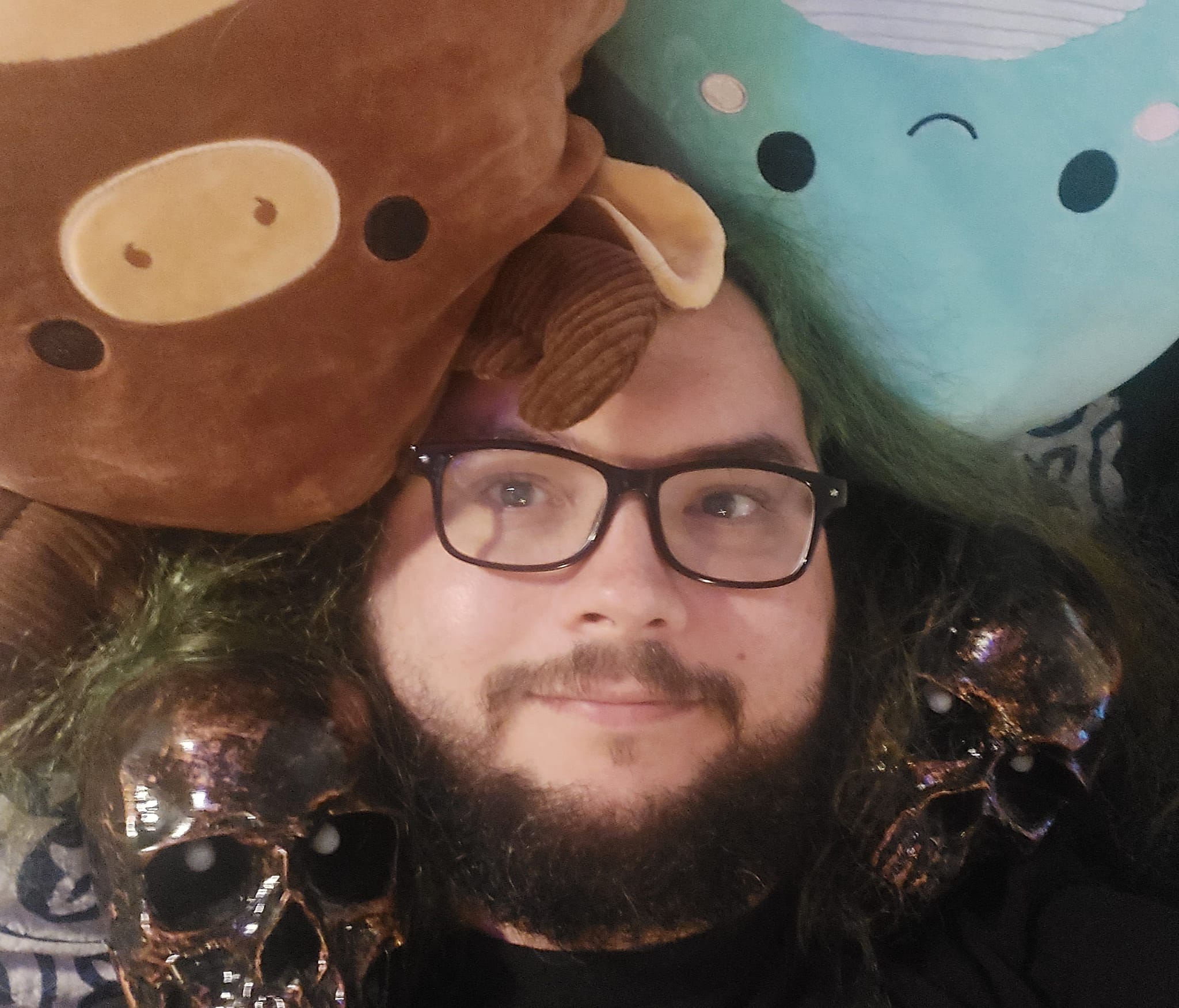 A man with green hair and a beard lays in bed with two plastic halloween skulls and two Squishmallows